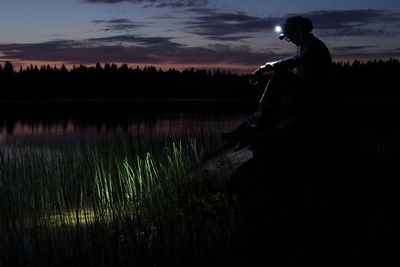 Silhouette man sitting with illuminated flashlight by lake against sky during sunset