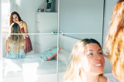 Smiling woman doing makeup for content female friend sitting on bed at home