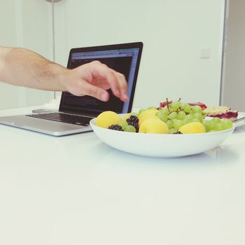 Cropped hand of man reaching out for fruits in bowl by laptop on table