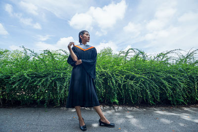 Woman wearing graduation gown standing on footpath against plants