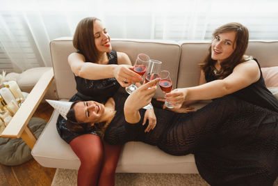 Cheerful friends holding drink sitting on sofa at home