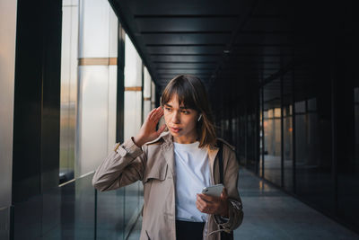 Serious female in stylish outfit and modern wireless earphones talking phone smartphone while standing near glass fence and modern building