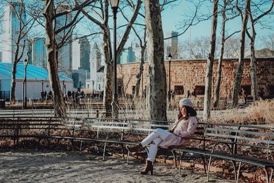 Woman sitting on seat against bare trees in a park