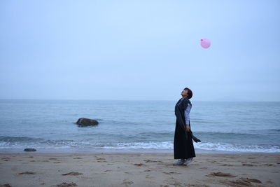 Young woman looking at balloon on shore against sky