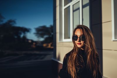 Young woman wearing sunglasses looking away while standing against built structure