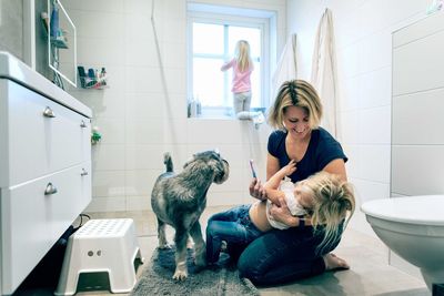 Smiling mother holding daughter with toothbrush by dog in bathroom