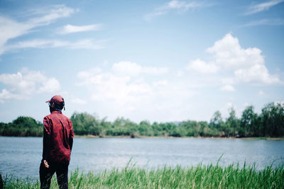 Rear view of man wearing cap standing by lake against sky