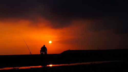 Silhouette man fishing on water against orange sky