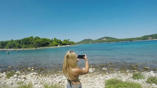 Rear view of woman photographing sea with smart phone against clear sky