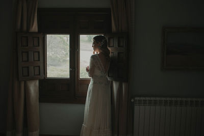 Side view of slim lady in white wedding dress with headband on hairs looking at window in room between darkness