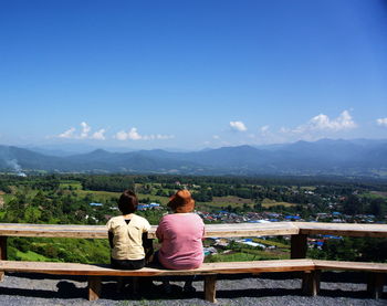 Rear view of friends looking at landscape while sitting on bench against blue sky