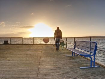 Traveler with backpack walk dificult with medicine poles. young tourist on bridge near sea at sunset