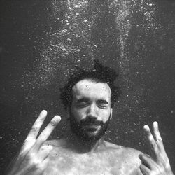 Shirtless man with eyes closed gesturing peace sign while swimming in sea