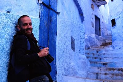 Portrait of happy man leaning on wall while holding digital camera