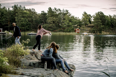 Daughters looking at parents fishing while sitting on rocky structure by lake