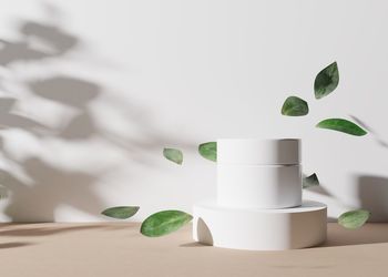 White and blank, unbranded cosmetic cream jar standing on white podium with leaves flying in the air