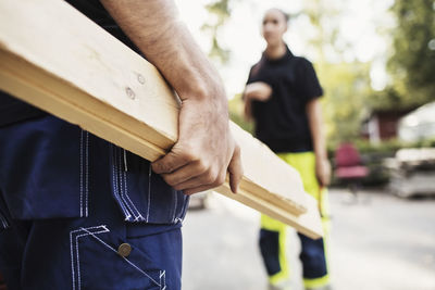 Midsection of male student carrying wooden planks with classmate standing in background