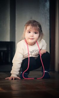 Cute girl with pink rope around neck crouching on hardwood floor at home