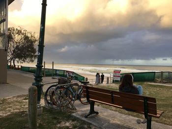 Rear view of woman sitting on bench by sea against cloudy sky