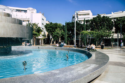 People sitting by fountain in city against sky