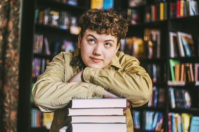 Thoughtful young man day dreaming while leaning on stack of books in bookstore