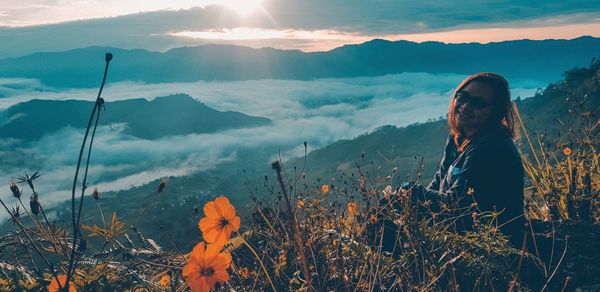 Portrait of smiling woman sitting on mountains against cloudy sky during sunset