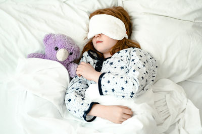 Girl  sleeps on a bed. she is wearing a sleep bandage over her eyes. next to her purple toy bear