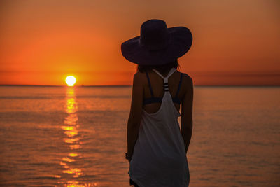 Rear view of woman standing by sea against sun during sunset