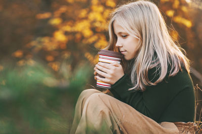 A beautiful teenage girl with blond hair sits thoughtfully in an autumn park, holds a thermos
