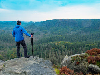 Photographer with folded tripod on end of cliff thinking. natural forestry landscape in valley below