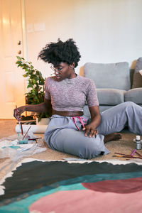 Young beautiful african american woman with afro hair, amateur artist in casual outfit drawing colorful picture with paintbrush on paper while sitting on floor in cozy apartment