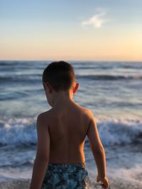 Rear view of shirtless boy at beach against sky during sunset