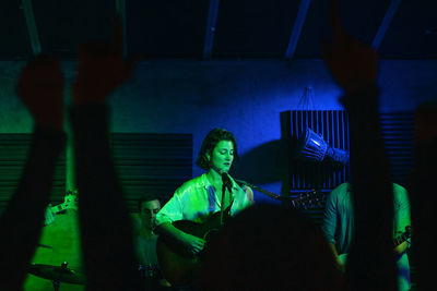 Group of people in casual clothes playing guitars and drums while woman singing and performing song in club with neon lights