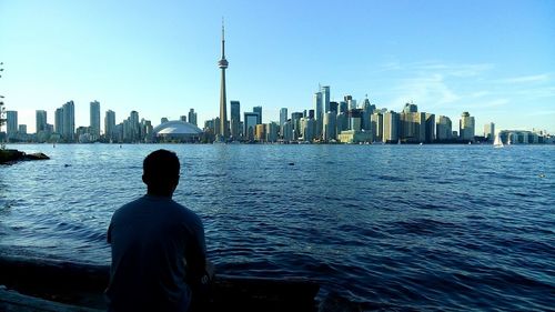 Rear view of man sitting by lake ontario against cn tower