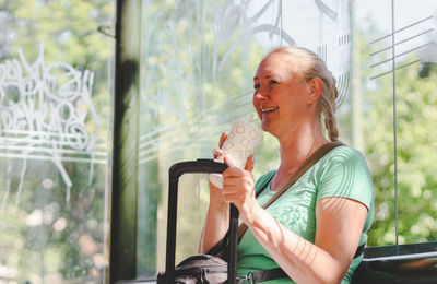 Young beautiful caucasian blonde woman sits on a bus stop bench outdoors holding a suitcase handle