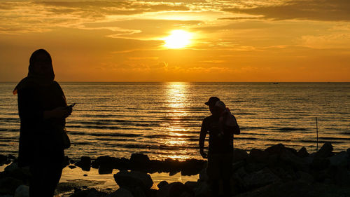 Silhouette people standing on rock by sea against sky during sunset