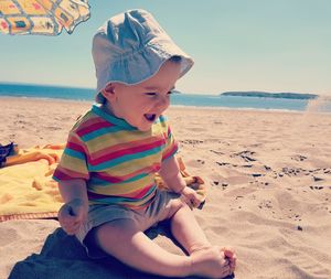 Happy toddler boy sitting on sand at beach against sky