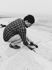 Side view of young man petting goat while crouching on sand