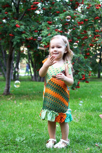Cute smiling little girl in colorful sundress is playing with soap bubbles in summer park.
