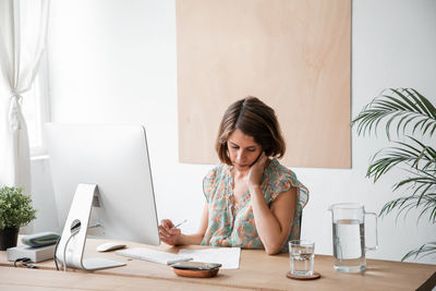 Concentrated female freelancer looking at document while sitting at desk with modern computer and water jug during remote work at home