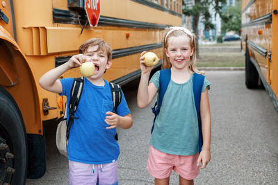 Children boy and girl students friends eating apples healthy snack by yellow school bus outdoors. 