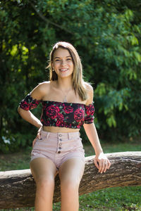 Portrait of smiling teenage girl sitting on tree trunk against plants in park