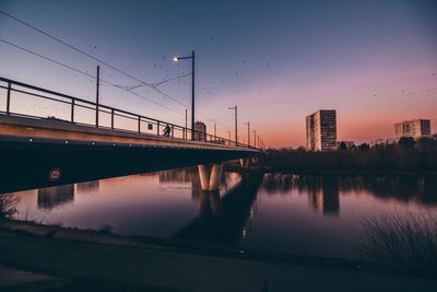 Bridge over river by buildings against sky at sunset