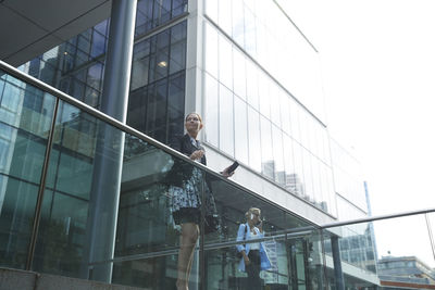 Businesswoman standing by railing with colleague talking on mobile phone in background at city