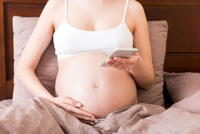 Caucasian pregnant girl using cellphone, resting on bed in home.