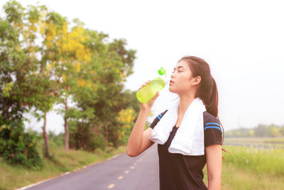 Young woman drinking water while standing against sky