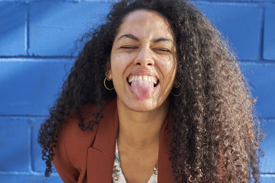 Woman sticking out tongue against blue wall on sunny day