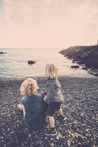 Rear view of mother with son sitting at beach against sky during sunset