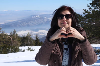 Portrait of smiling mature woman making heart shape while standing on snow field