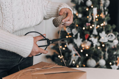 Midsection of woman wrapping christmas present in kraft paper in front of christmas tree at home